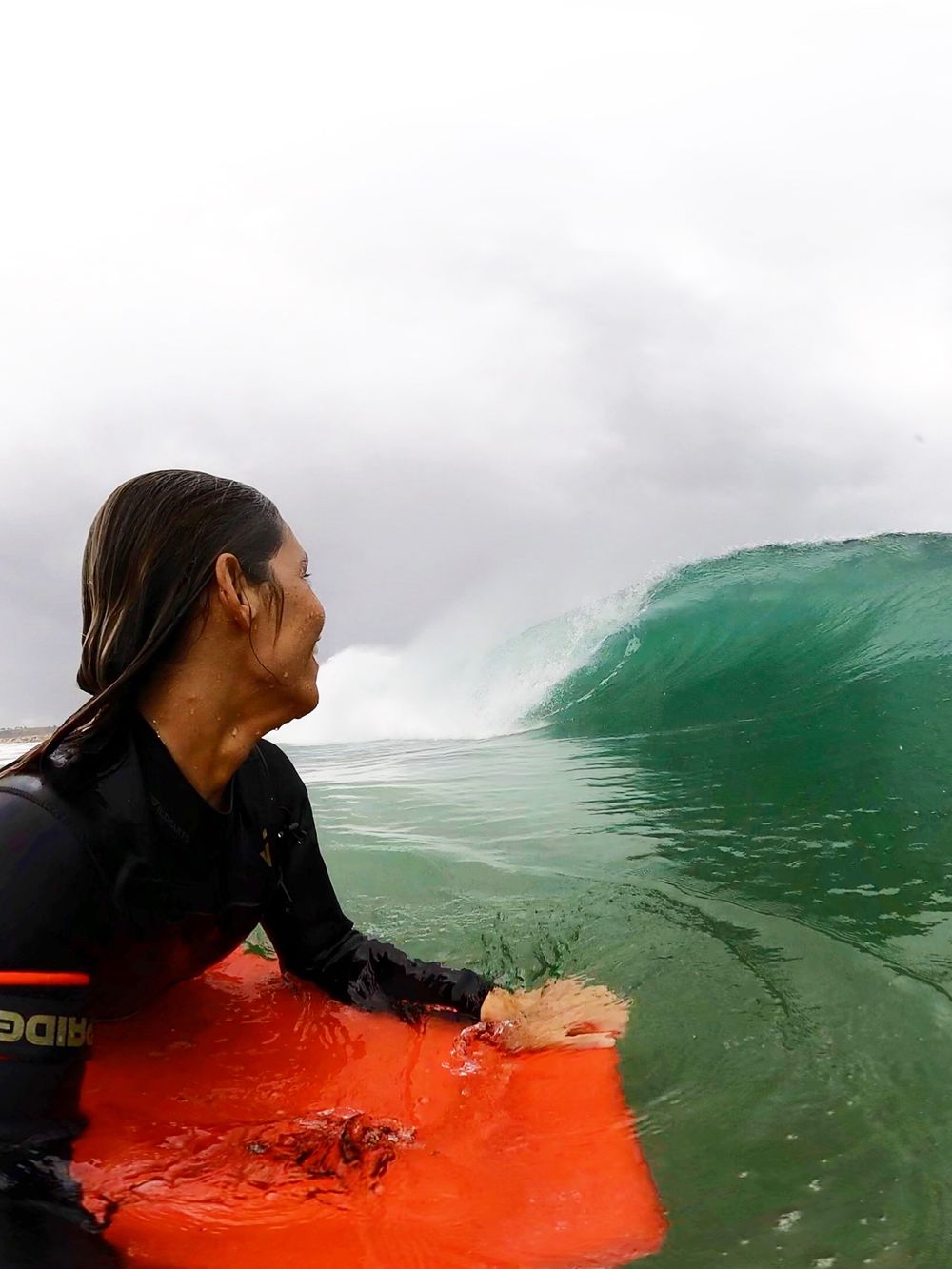 Isabela Sousa na água, ao lado da prancha laranja, observando a onda se formar.